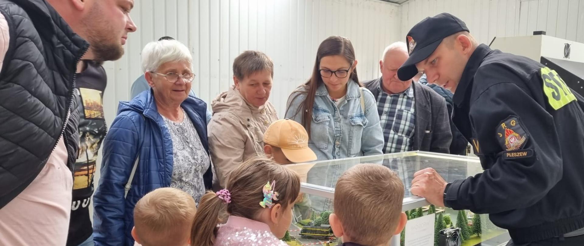 Grupa dzieci i dorosłych stojących przed stoiskiem edukacyjnym PSP, widoczny mobilny symulator zagrożeń z modułem „jezioro – las”