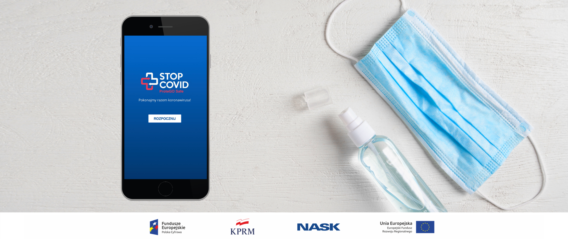 Z prawej strony - leżące na białym tle maseczka chirurgiczna (widoczna niebieską stroną) i przezroczyste opakowanie z płynem do dezynfekcji. Po lewej stronie czarny telefon z ekranem startowym aplikacji STOP COVID - ProteGO Safe.