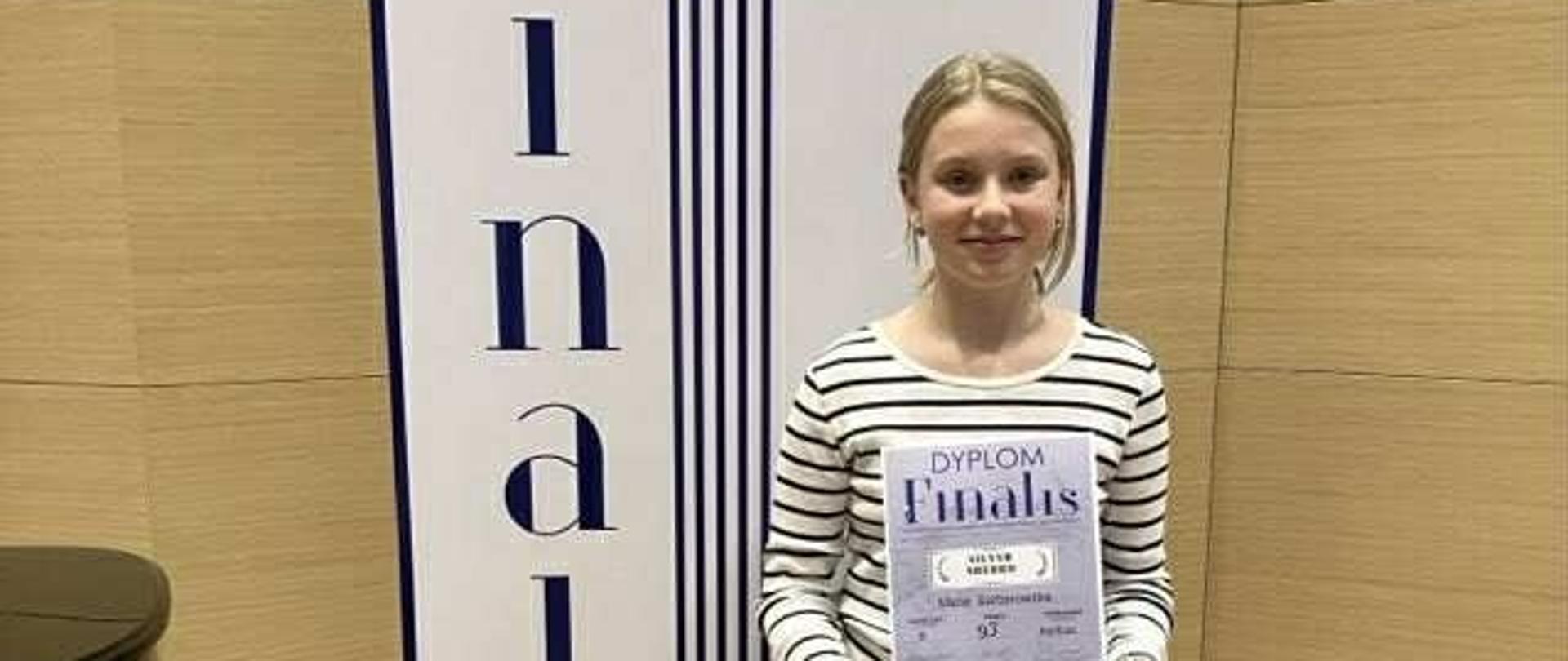 Uczennica stoi przed ścianką reklamową konkursu i trzyma dyplom. 