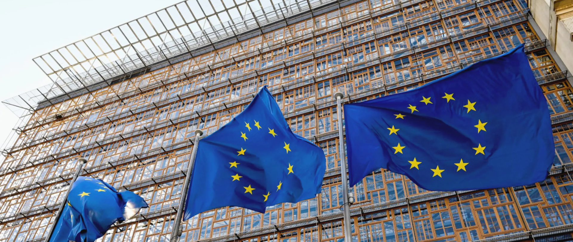 Trzy flagi Unii Europejskiej wiszą na masztach, w tle budynek biurowy.