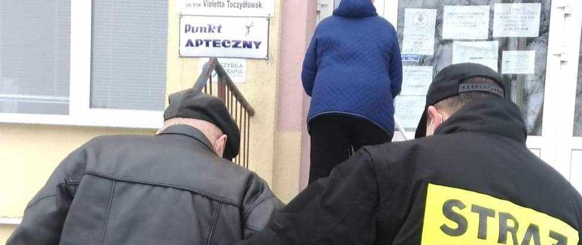 Na zdjęciu strażak OSP w ubraniu koszarowym pomaga wejść po schodach starszej osobie do przychodni NFZ.