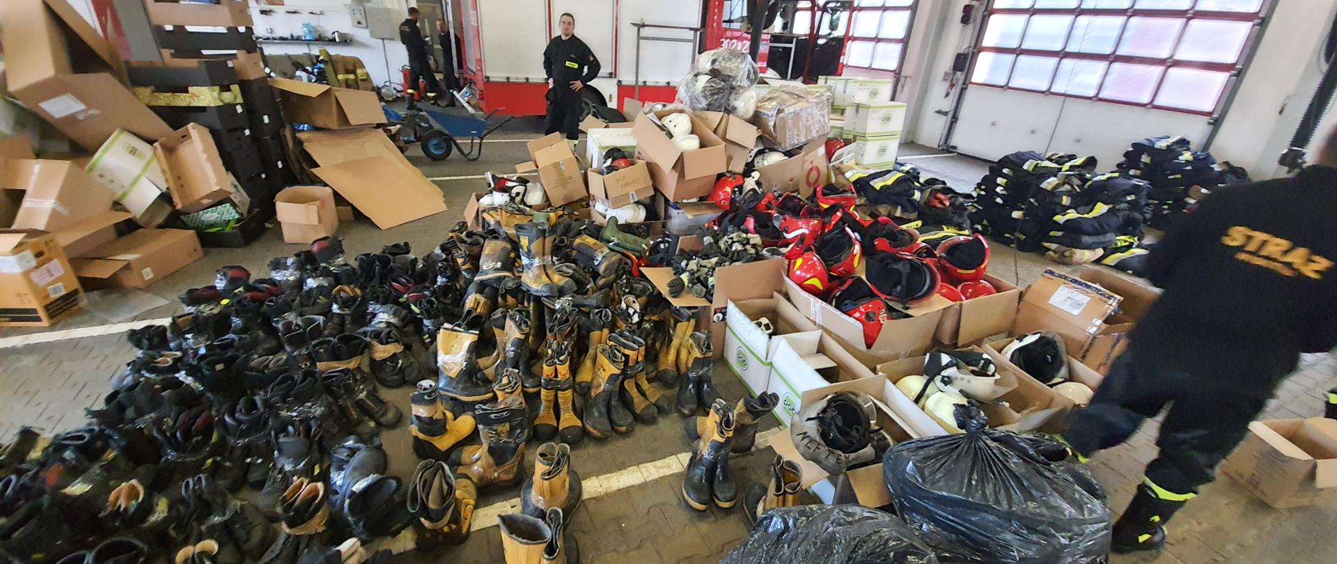na zdjęciu dwóch strażaków oraz bardzo duże ilości butów hełmów i ubrań strażackich w tle samochód gaśniczych, zdjęcie zrobione w garażu jednostki ratowniczo gaśniczej