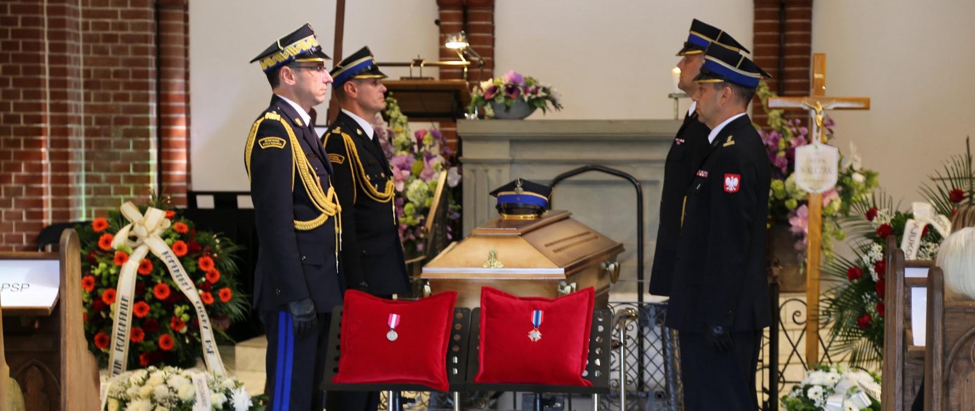 Zdjęcie przedstawia wnętrze kościoła lub kaplicy, gdzie odbywa się ceremonia pogrzebowa. W centrum, na katafalku, znajduje się jasna trumna ozdobiona czapką z czarnym daszkiem i złotym emblematem.
Trumnę otaczają czterej mężczyźni w galowych mundurach Państwowej Straży Pożarnej. Mundury są granatowe, ze złotymi guzikami i insygniami. Mężczyźni stoją na baczność, twarze mają poważne, a ich pozycja sugeruje oddanie hołdu zmarłemu.
Przed trumną, na dwóch oddzielnych stojakach, leżą dwie czerwone poduszki. Na każdej z nich umieszczony jest okrągły, srebrny medal. Na poduszkach widać też białe sznurki z chwostami.
Wokół trumny i mężczyzn widać wiele kwiatów i wieńców pogrzebowych w kolorach białym, czerwonym, różowym i fioletowym. Z prawej strony widać fragment ołtarza, a na ścianie w tle wisi drewniany krzyż. Na podłodze ułożone są jasne, kwadratowe płytki. W tle widać ceglaną ścianę i drewniane ławki, co wskazuje na sakralne wnętrze. Całość kompozycji jest uroczysta i pełna szacunku.
