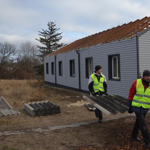 Dwóch mężczyzn niesie fragment dachówki. Ubrani są w kamizelki z logo Polska pomoc