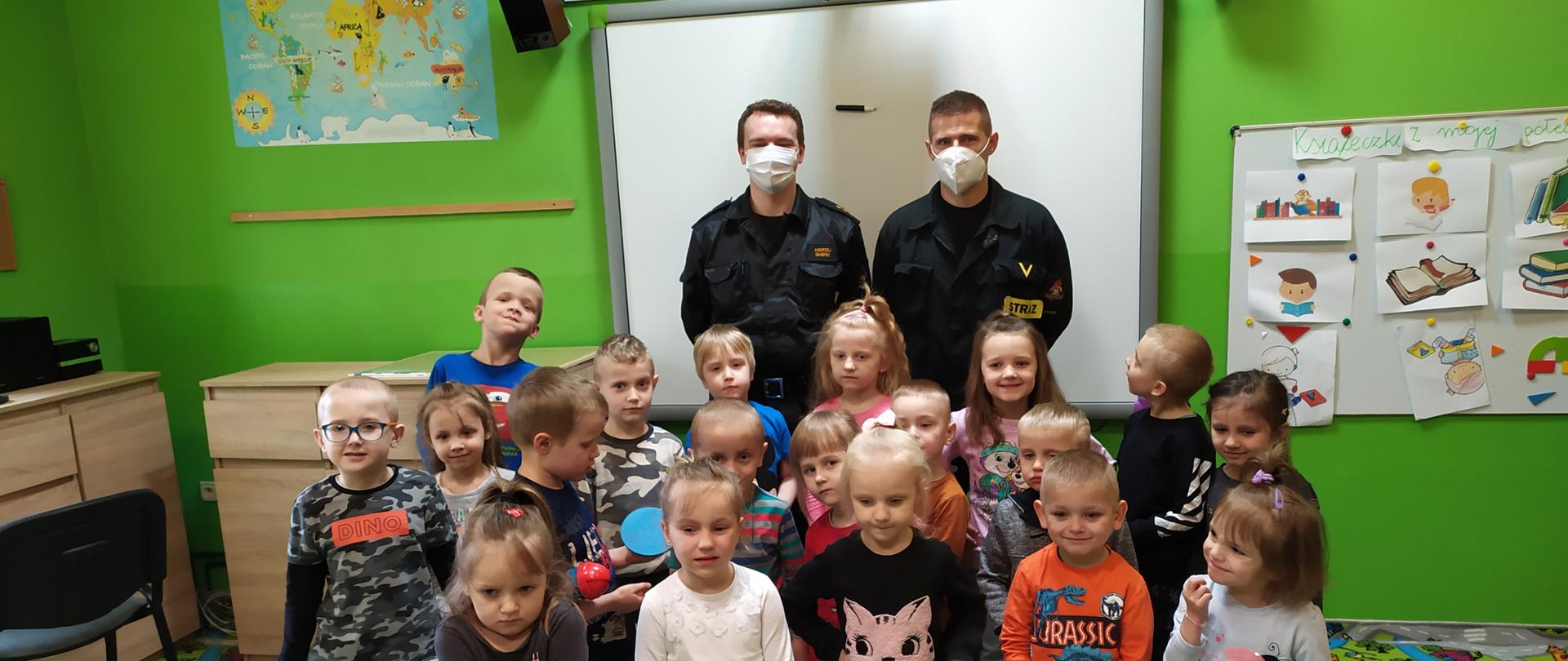 Na zdjęciu widać grupę dzieci w wieku przedszkolnym. Za grupą dzieci stoją dwaj funkcjonariusze PSP ubrani w czarne mundury. Ściany pomieszczenia są w kolorze zielonym. Osoby stoją na dywanie na którym jest plan miasta. Obok grupy osób, po prawej stronie rysunki dzieci na ścianie. W tle widać białą tablicę, nad głowami rzutnik. Po lewej stronie brązowe umeblowanie Sali, krzesło. Na ścianie mapa edukacyjna.