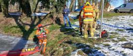 Na brzegu akwenu stoją strażacy przygotowujący się do akcji. Na lodzie znajdują się sanie lodowe. W tle widoczny jest park, zabudowania i zaparkowane samochody.