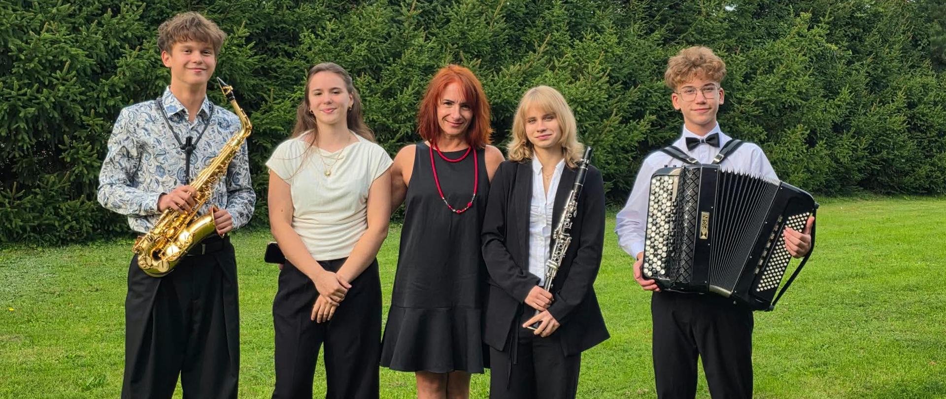 Grupa uczniów wraz z akompaniatorką (w środku) pozuje na trawniku na tle zielonych iglaków. Młodzi muzycy trzymają swoje instrumenty – od lewej widoczny jest saksofon, następnie klarnet oraz akordeon. Wszyscy są elegancko ubrani i uśmiechają się w stronę obiektywu.