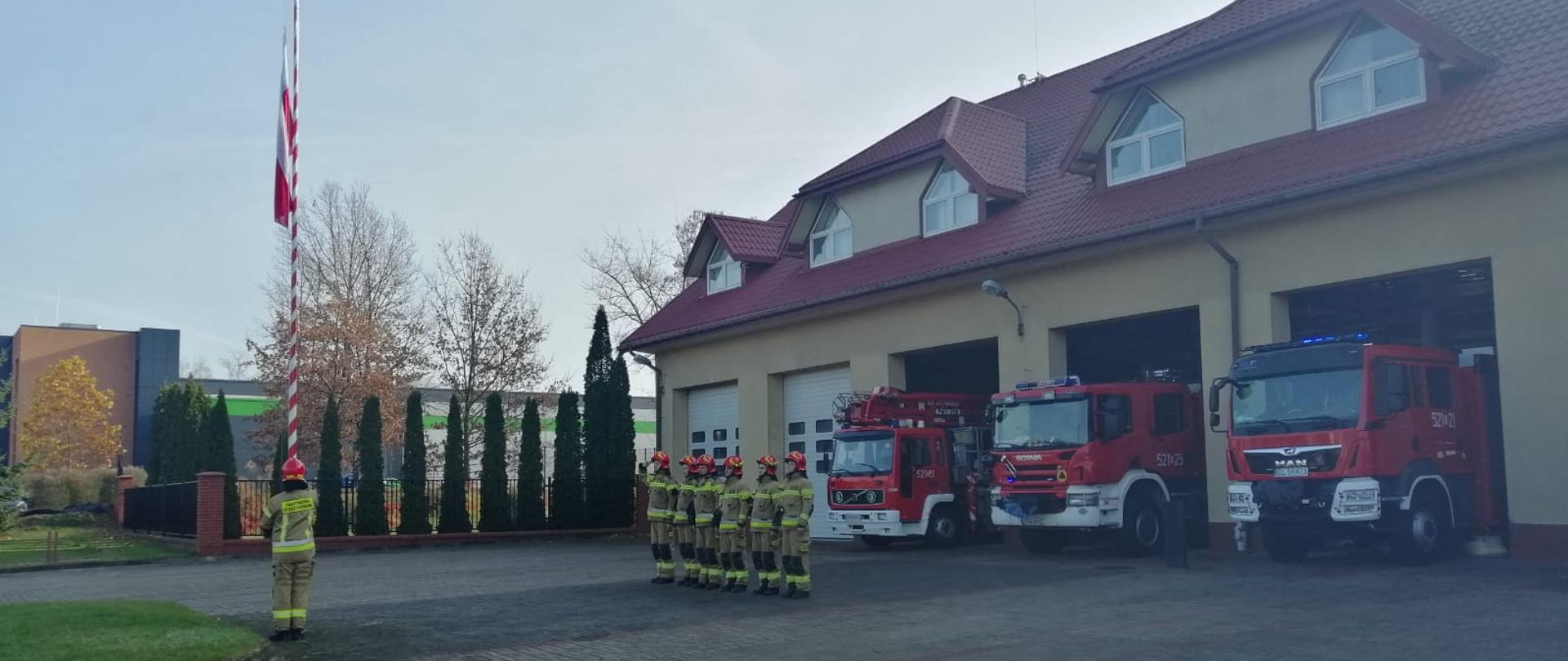 Na zdjęciu widać 7 strażaków. 6 z nich stoi w szeregu, zwróconych twarzami do masztu na którą wciągana jest flaga Polski przez 1 strażaka. Za strażakami ustawionymi w szeregu znajduję się strażnica z 3 samochodami strażackimi stojącymi w otwartych bramach garażowych. Strażacy ubrani są w ubrania bojowe w kolorze piaskowym i czerwone hełmy. 