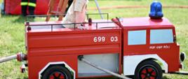 Pomniejszony wóz strażacki w kolorze czerwonym stoi na trawie. Z tyłu i z boku wystają z niego węże strażackie w kolorze białym. Na dachu wozu stoi dziecko w czapce z daszkiem. W tle widać zbiornik na wodę oraz stół z ławkami.