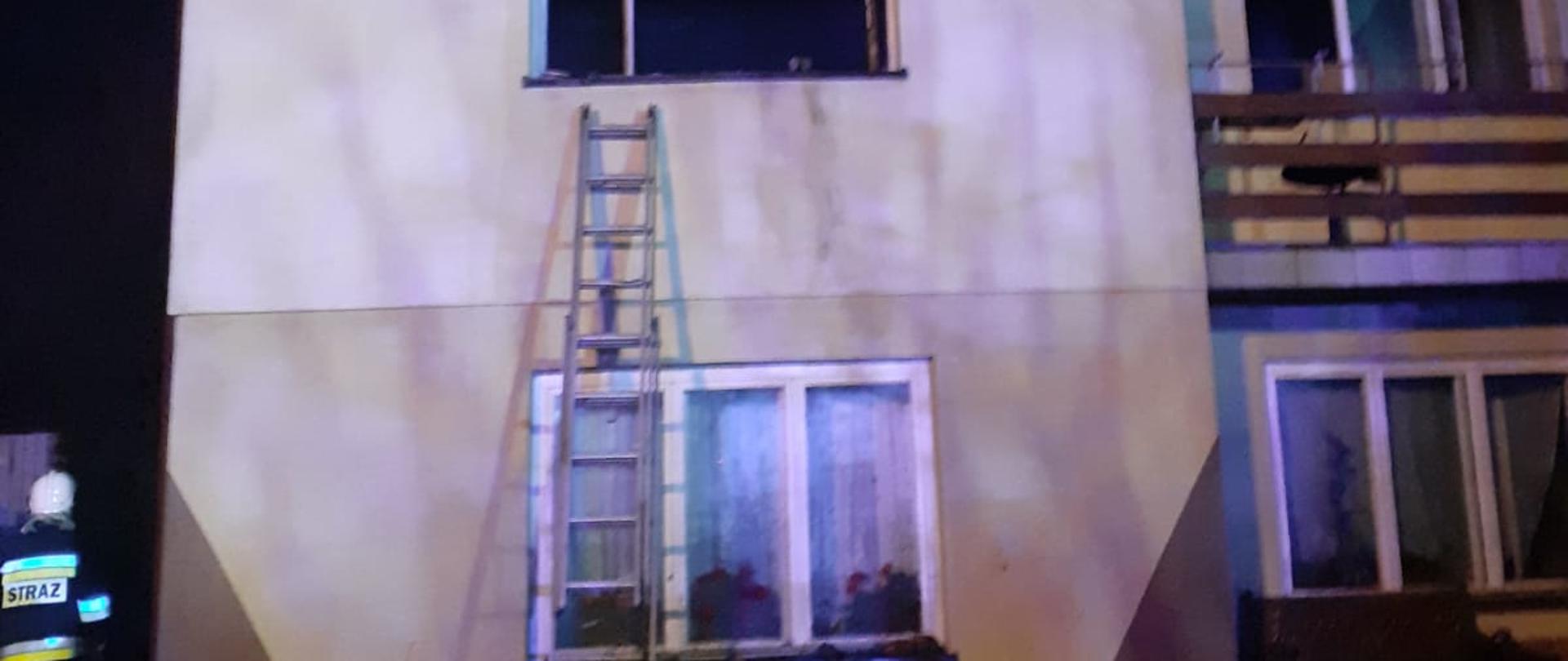 Na zdjęciu widoczny budynek mieszkalny wielokondygnacyjny oraz przystawiona drabina do okna na I piętrze. Z lewej strony obok budynku widoczni strażacy. U dołu zdjęcia znajdują się spalone elementy wyposażenia wnętrza.