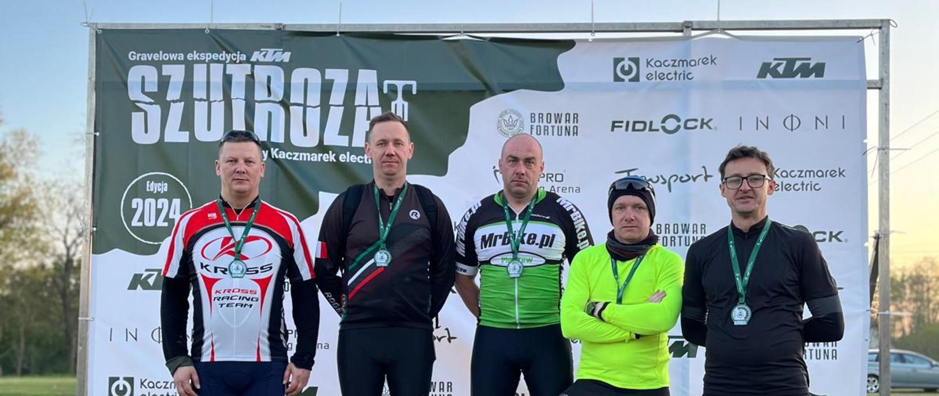 Na podium stoi pięciu mężczyzn ubranych w stroje sportowe, każdy z nich ma na sobie medal. Za nimi widoczny baner z zielonym logiem imoprezy sportowej szutroza