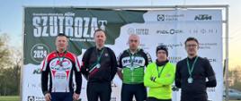 Na podium stoi pięciu mężczyzn ubranych w stroje sportowe, każdy z nich ma na sobie medal. Za nimi widoczny baner z zielonym logiem imoprezy sportowej szutroza