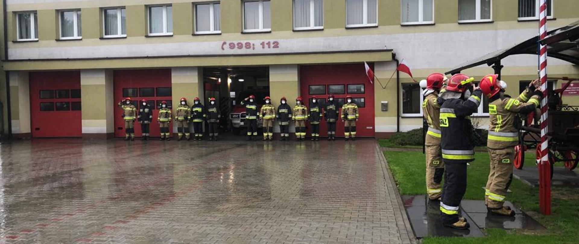 Zdjęcie przedstawia poczet flagowy złożony z trzech strażaków zawieszający flagę na maszcie na tle budynku Komendy Powiatowej Państwowej Straży Pożarnej w Wieluniu i strażaków zmian służbowych