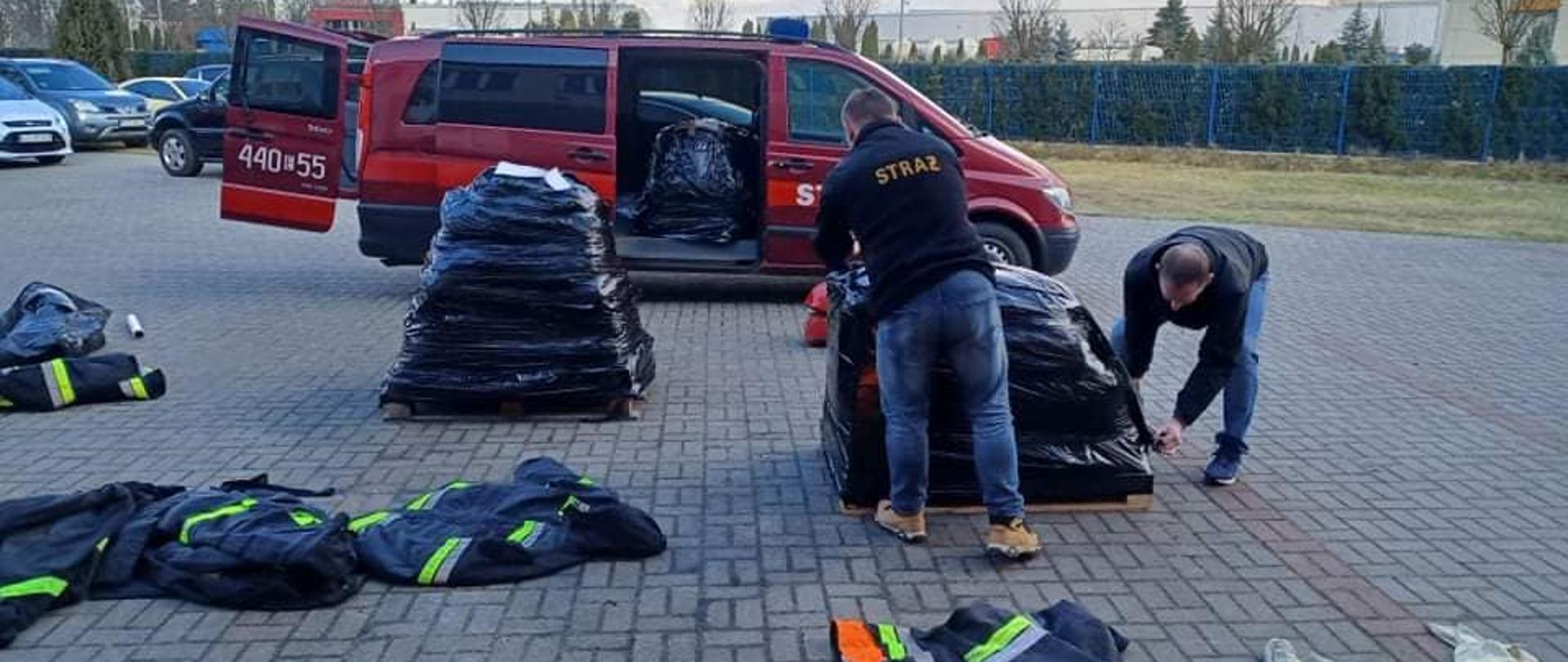 Na zdjęciu widać dwóch mężczyzna, którzy obwijają czarną folią paletę. Obok stoi druga już obwinięta paleta czarną folią. Na kostce brukowej leżą ubrania strażackie gotowe do spakowania. W tle widać czerwony samochód strażacki typu bus, do którego pakowane są palety. W tle widać drzewa i samochody osobowe. Jest widno. 