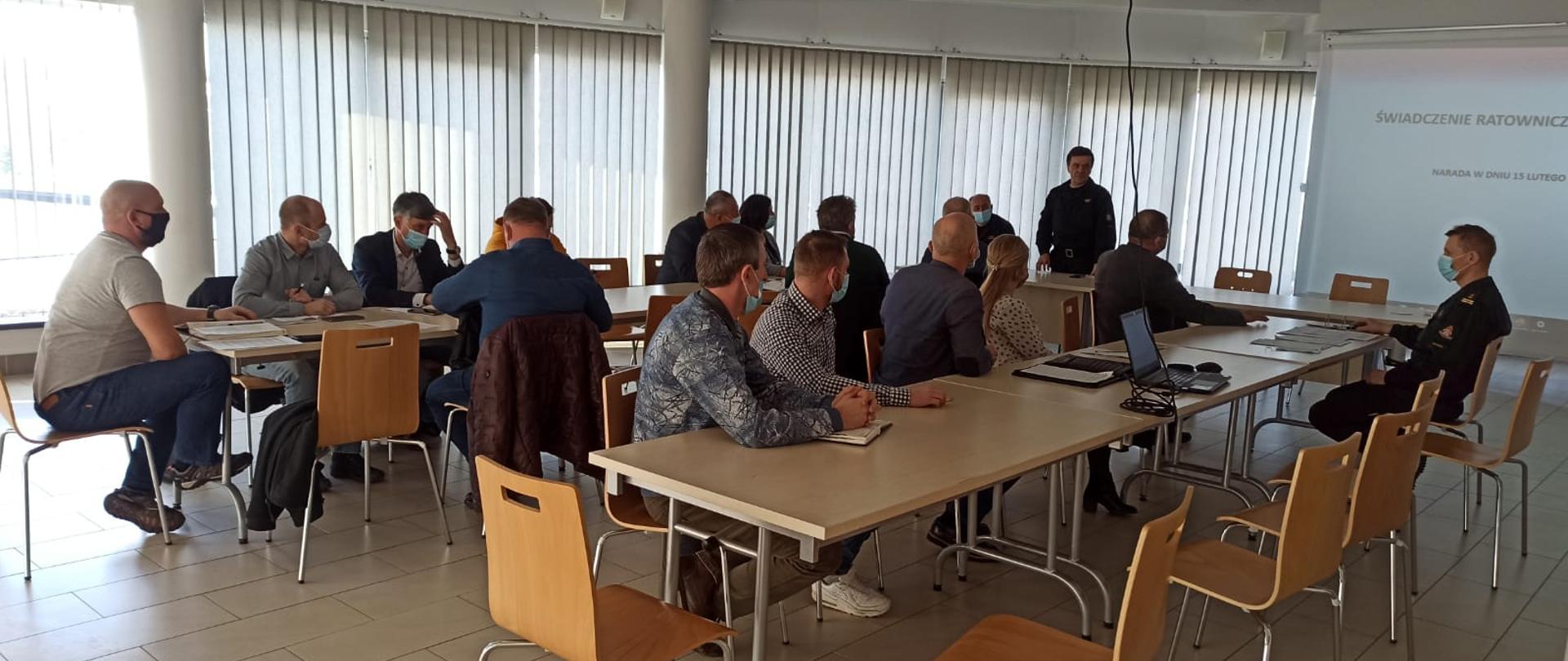 Zdjęcie przedstawia pracowników gminy oraz strażaków, którzy siedzą przy stolikach i słuchają wystąpienia komendanta powiatowego dotyczącego ustawy o OSP. Na drugim planie widać rzutnik i przedstawianą prezentację