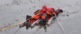 Strażak w skafandrze wypornościowym przy użyciu dwóch przęseł drabiny nasadkowej dociera do osoby poszkodowanej znajdującej się w przeręblu celem jej ewakuacji z akwenu zalodzonego . 