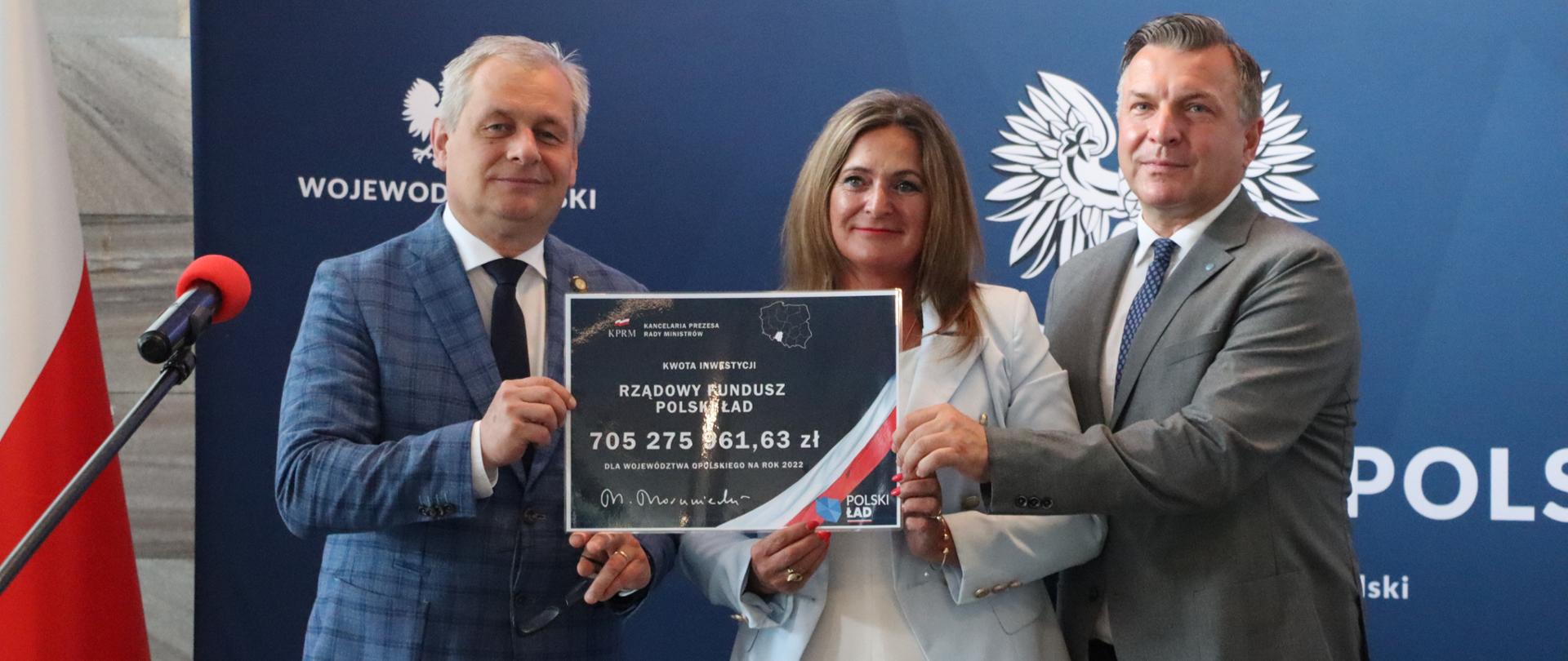 Na zdjęciu wojewoda opolski, poseł Czochara i pracownik Biura Wojewody, trzymają oni wydrukowany czek z kwotą 705 mln zł dla regionu. W tle ścianka z napisem Wojewoda Opolski. Po lewej stronie biało-czerwona flaga. 