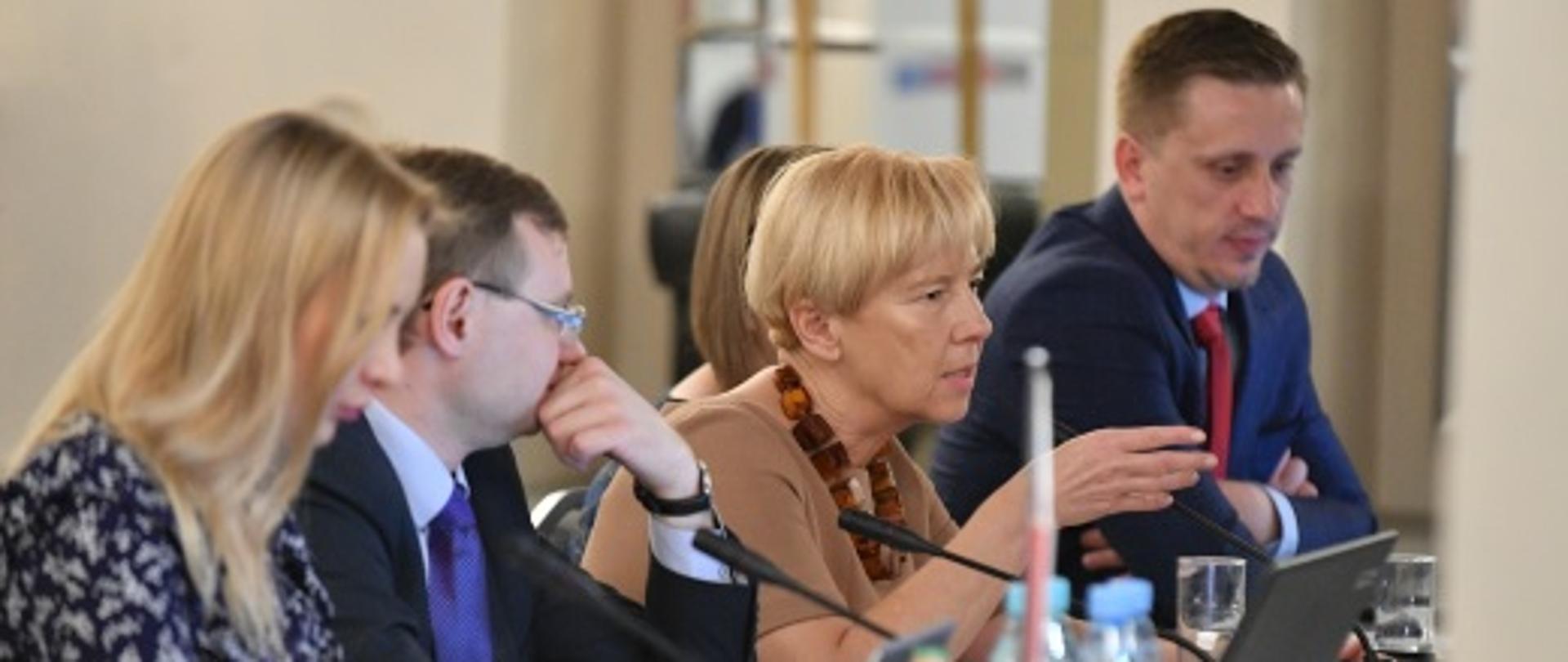 Minister Zielińska siedzi przy stole na którym stoją laptopy i mikrofony. Obok niej znajdują się pozostali uczestnicy debaty. 