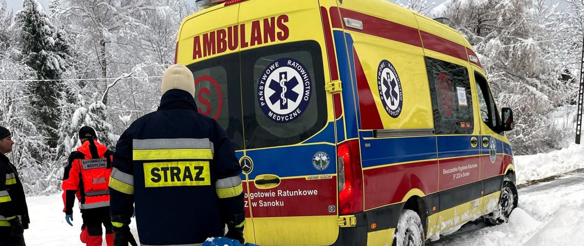 Zdjęcie przedstawia karetkę pogotowia która stoi na drodze za nią stoi dwóch strażaków OSP oraz jeden ratownik medyczny. 