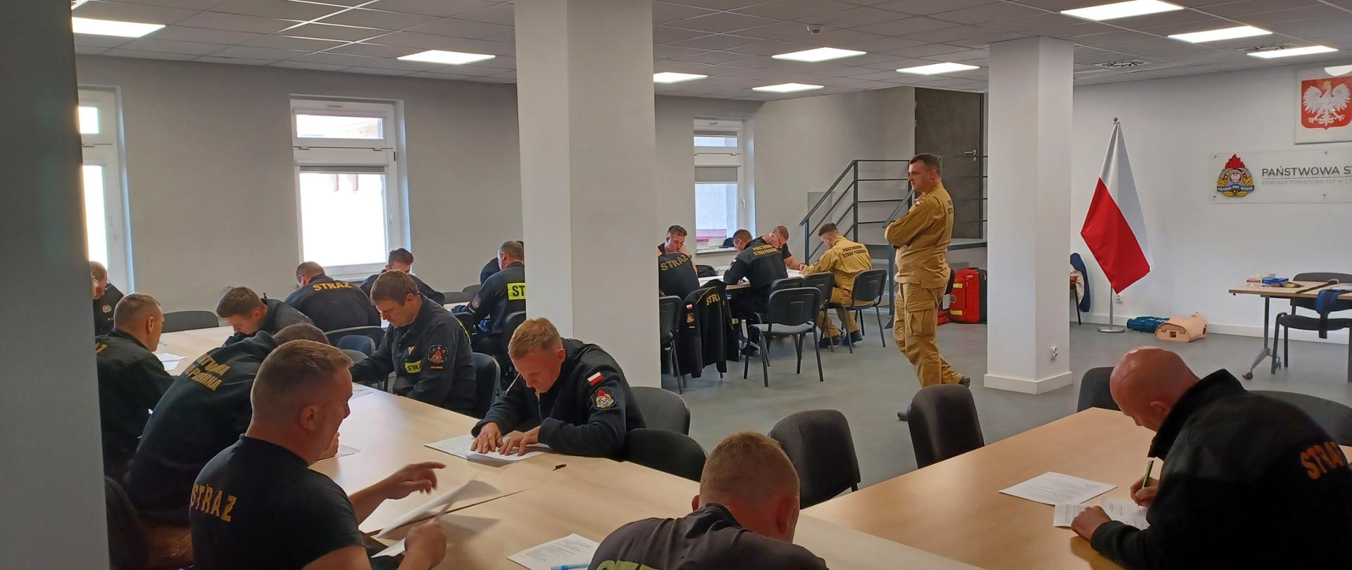 Strażacy siedzą przy stole na sali w Komendzie Powiatowej Państwowej Straży Pożarnej w Człuchowie i rozwiązują test części teoretycznej egzaminu. Na środku sali stoi przewodniczący komisji egzaminacyjnej.