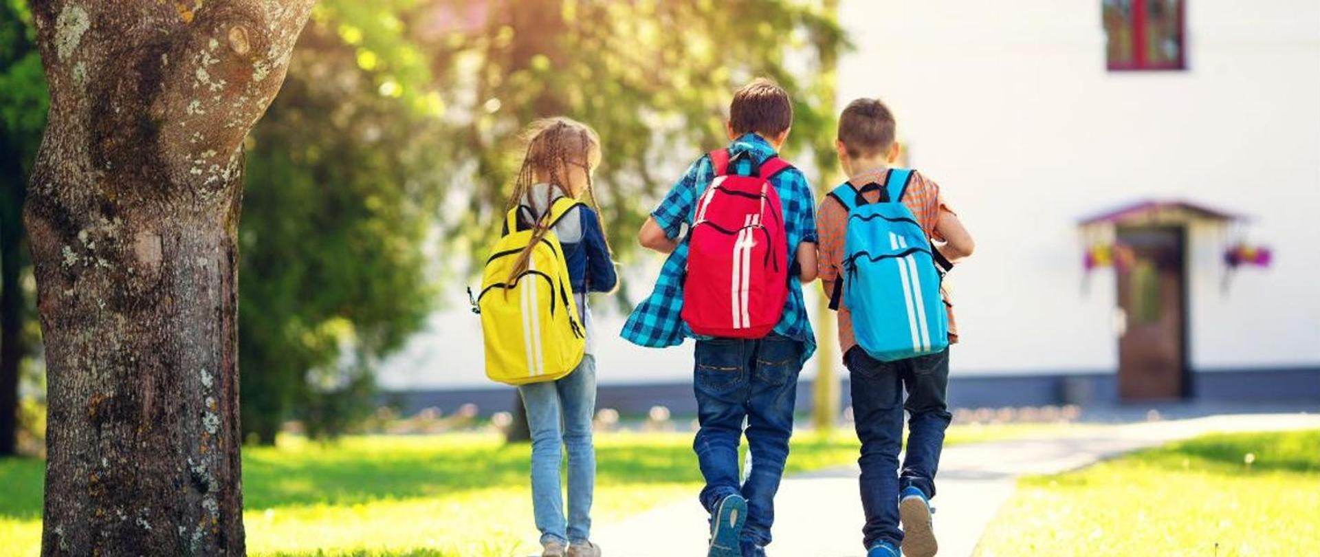 Trójka dzieci: dziewczynka z żółtym tornistrem i dwójka chłopców z czerwonym i niebieskim tornistrem idą ścieżką do szkoły.