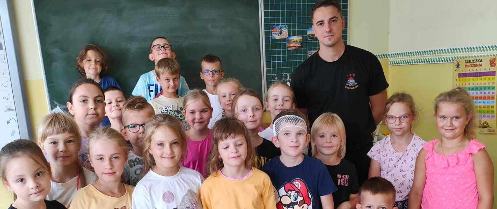 Zdjęcie przedstawia Strażaka i dzieci stojące przy nim pozując do wspólnego zdjęcia. Wszyscy stoją na tle tablicy szkolnej a przed nimi widoczny jest spakowany w niebieskie torby sprzęt szkoleniowy.