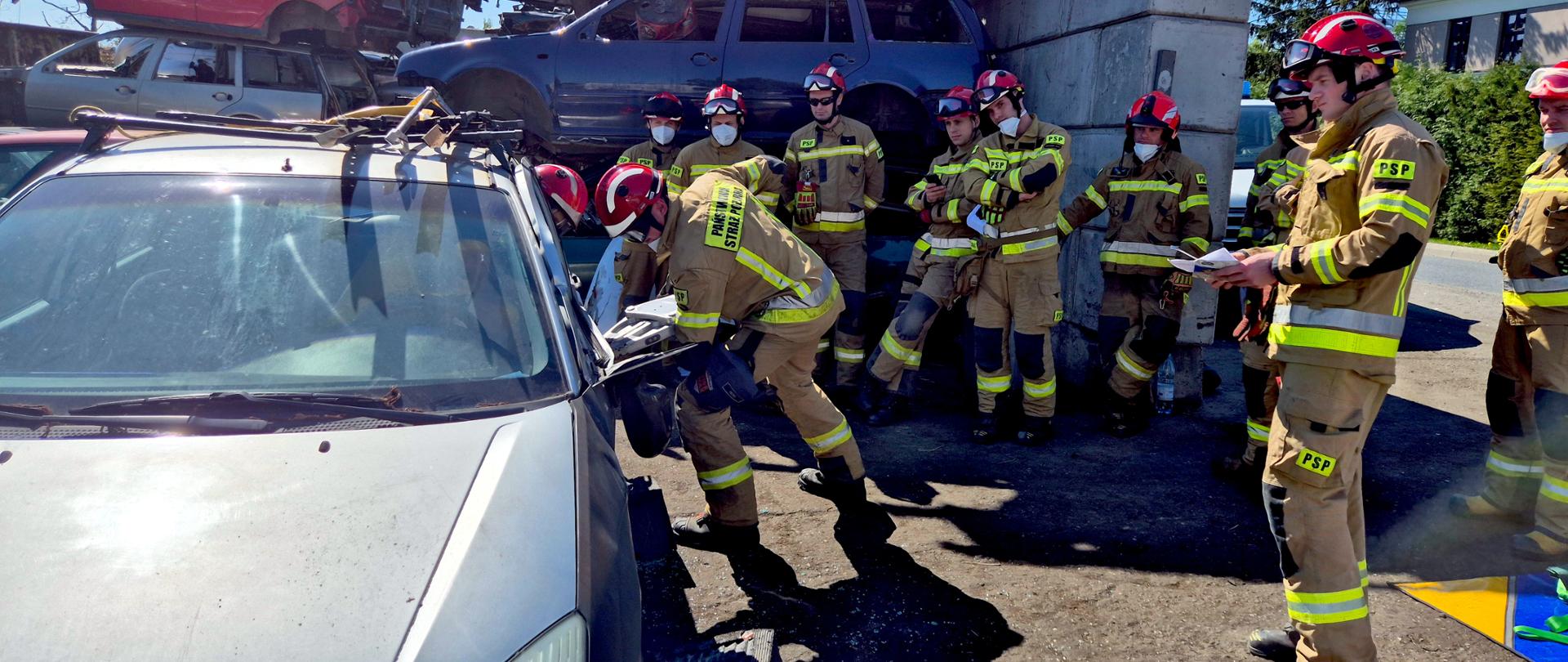 Na zdjęciu grupa strażaków w jasnożółtych ubraniu specjalnym, ochronnym z odblaskowymi paskami i czerwonymi hełmami uczestniczy w ćwiczeniach ratownictwa technicznego na złomowisku samochodowym. Na ich mundurach widoczne są żółte naszywki z czarnymi literami „PSP” (Państwowa Straż Pożarna). W tle widać stos starych, zniszczonych samochodów ułożonych jeden na drugim oraz betonowe bloki zabezpieczające przestrzeń.
Na pierwszym planie jeden strażak pochyla się nad wrakiem srebrnego samochodu, używając narzędzi do jego rozcinania lub rozginania karoserii — ćwiczy techniki ratownictwa drogowego. Pozostali strażacy uważnie obserwują jego działania, a jeden z nich trzyma w rękach notatnik lub instrukcję. Wszyscy wyglądają na skupionych i zaangażowanych.
