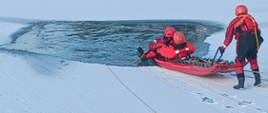 Na skraju przerębla troje strażaków ćwiczy wyciąganie z wody osoby poszkodowanej. Strażacy ubrani są w ubrania koloru czerwonego do pracy w wodzie. Do wyciągnięcia służą im sanie lodowe. Ratownicy zabezpieczeni linkami asekuracyjnymi oraz kapokami.