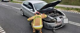 Zdjęcie przedstawia uszkodzony pojazd marki Alfa Romeo w obrębie, którego działania realizuje strażak PSP. 
