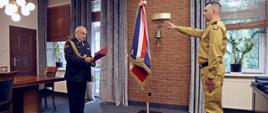 Gabinet Komendanta Centralnej Szkoły PSP, po prawej stojący na baczność w mundurze koloru piaskowego ślubujący funkcjonariusz, przed nim stojący w mundurze galowym ze sznurem Komendant Centralnej Szkoły PSP odczytujący z trzymanej w dłoniach czerwonej teczki tekst ślubowania. Pomiędzy nimi stojący na stojaku sztandar Szkoły