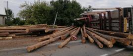 Na zdjęciu widać wywrócony na drogę samochód ciężarowy przewożący dłużycę drewna. Na drodze widać dłużycę drewna.