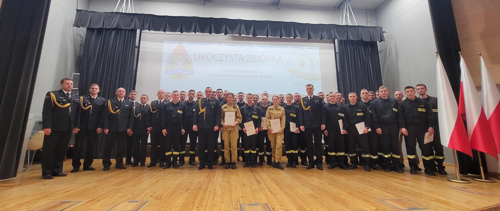 Pamiątkowe zdjęcie uczestników szkolenia podstawowego SP1/24. Na scenie auli SA PSP w Krakowie ubrani w umundurowanie wyjściowe, służbowe i koszarowe strażaczki i strażacy. Po prawej stronie stojak z biało-czerwonymi flagami. Za grupą wyświetlany na białym tle okolicznościowy slajd. 