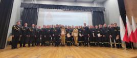 Pamiątkowe zdjęcie uczestników szkolenia podstawowego SP1/24. Na scenie auli SA PSP w Krakowie ubrani w umundurowanie wyjściowe, służbowe i koszarowe strażaczki i strażacy. Po prawej stronie stojak z biało-czerwonymi flagami. Za grupą wyświetlany na białym tle okolicznościowy slajd.