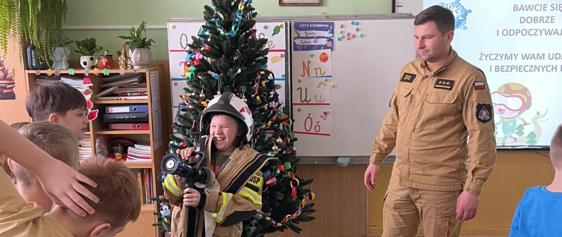 Bezpieczne ferie 2025 strażak pokazuje sprzęt będący na wyposażeniu KP PSP w Lipsku. Zdjęcie przedstawia zajęcia edukacyjne z udziałem strażaków w szkolnej klasie. W centrum kadru, po prawej stronie, stoi dorosły mężczyzna w mundurze strażackim, uśmiechający się i patrzący na dzieci.
Na pierwszym planie, w środku, widoczna jest dziewczynka, ubrana w zbyt duży jak na nią mundur strażacki i hełm, z szerokim uśmiechem na twarzy, trzymająca w rękach prądownicę. Obok niej i za nią widać inne dzieci, które z zaciekawieniem obserwują scenę. W tle, za dziewczynką, stoi choinka udekorowana bombkami, a za mężczyzną na białej tablicy wiszą pomoce naukowe, w tym "Lista Dyżurnych" i plansza z literami. Na tablicy po prawej stronie widnieje hasło "BAWCIE SIĘ DOBRZE I ODPOCZYWAJĄ" oraz "ŻYCZYMY WAM UDANYCH I BEZPIECZNYCH". Klasa jest dobrze oświetlona.