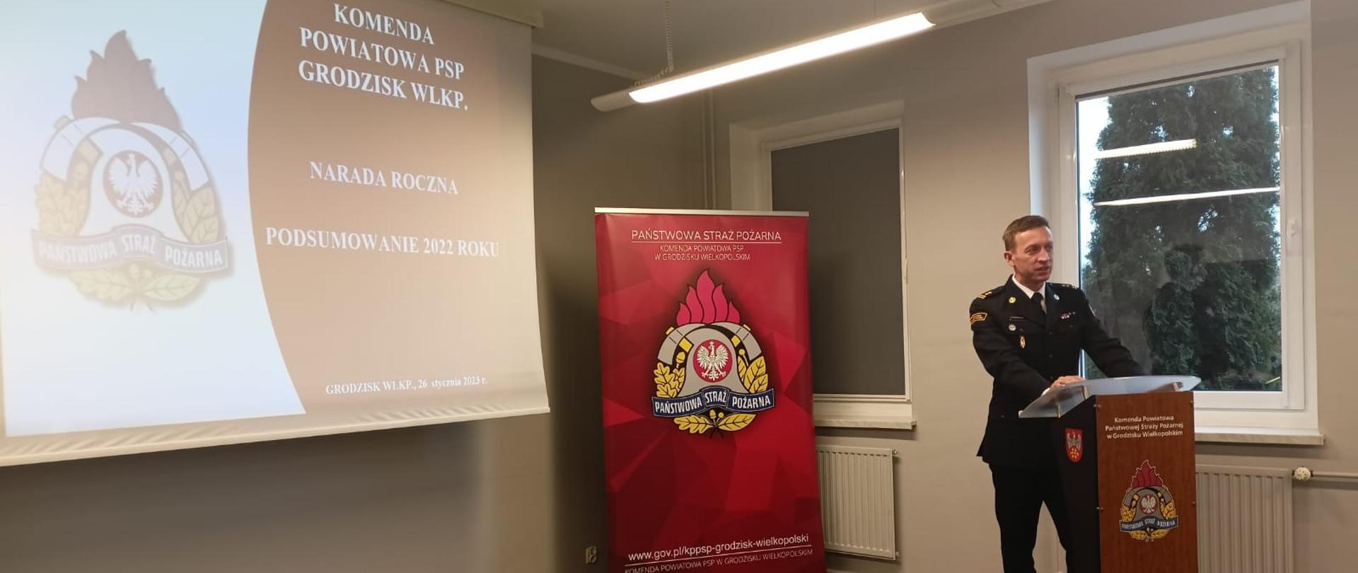 strażak w mundurze wyjściowym stoi przed mównicą, w tle duży ekran z logo PSP oraz czerwony baner z logo psp