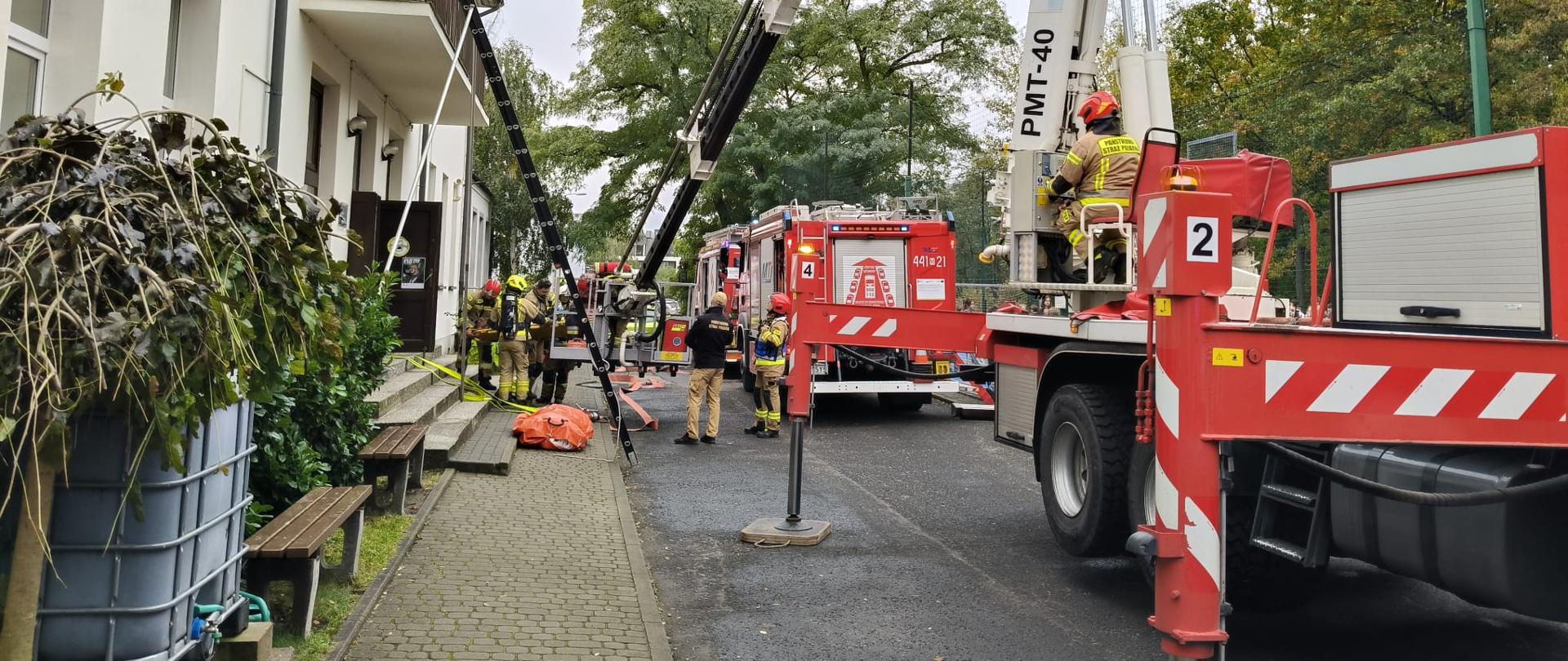 jęcie przedstawia scenę z ćwiczeń ratowniczych straży pożarnej na terenie przed białym, dwupiętrowym budynkiem (prawdopodobnie szkołą). W centrum uwagi znajduje się czerwony wóz strażacki oraz operujący podnośnik koszowy. Kilku strażaków w ubraniach specjalnych i hełmach wykonuje czynności przy sprzęcie. Na pierwszym planie widoczna jest duża, rozstawiona drabina, pomarańczowy pokrowiec ratowniczy oraz żółte węże pożarnicze. Tło stanowią drzewa o jesiennym listowiu.