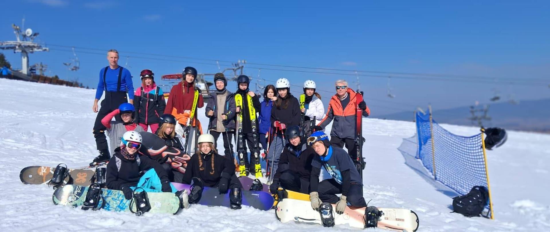 Grupa młodzieży i opiekunów pozuje do wspólnego zdjęcia na stoku narciarskim pod bezchmurnym, niebieskim niebem.
Większość osób ma na sobie kaski i stroje narciarskie; część siedzi na śniegu z deskami snowboardowymi, a inni stoją z nartami w dłoniach. W tle widoczny jest wyciąg krzesełkowy oraz siatka zabezpieczająca trasę.