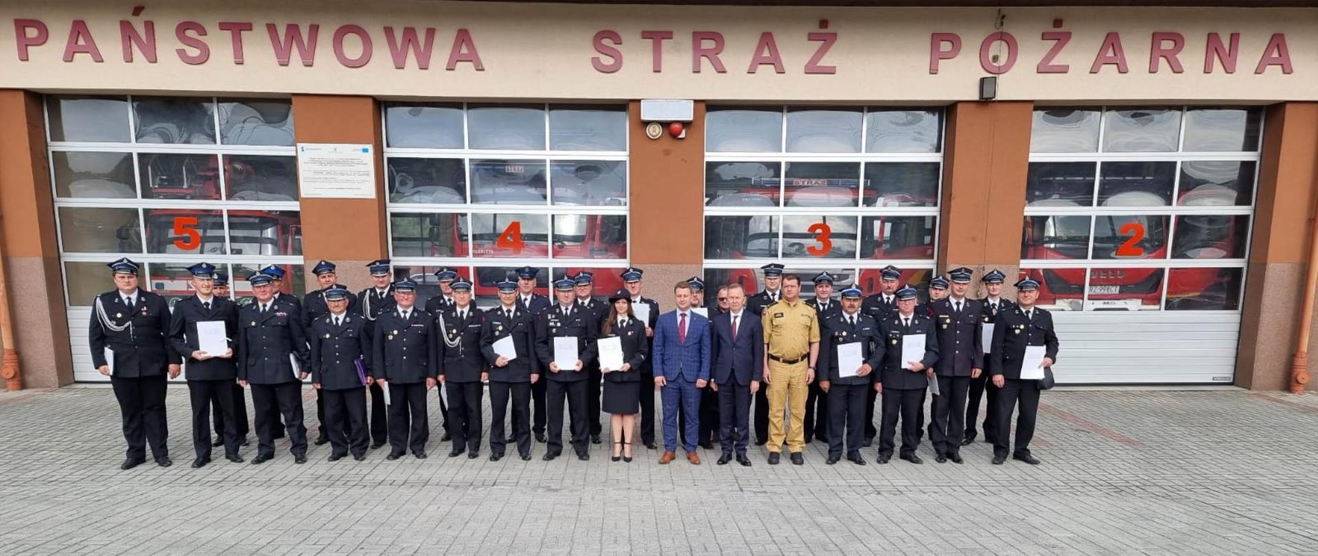 Na placu przed garażami stoi grupa osób. Większość to strażacy OSP. Za nimi widoczne bramy garażowe i Napis Państwowa Straż Pożarna.