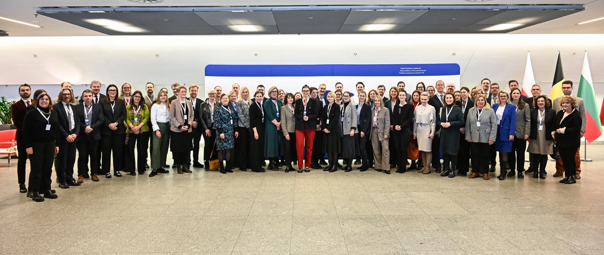 Zdjęcie zbiorowe, pod białą ścianą na szarej podłodze stoi duża grupa kilkudziesięciu osób.