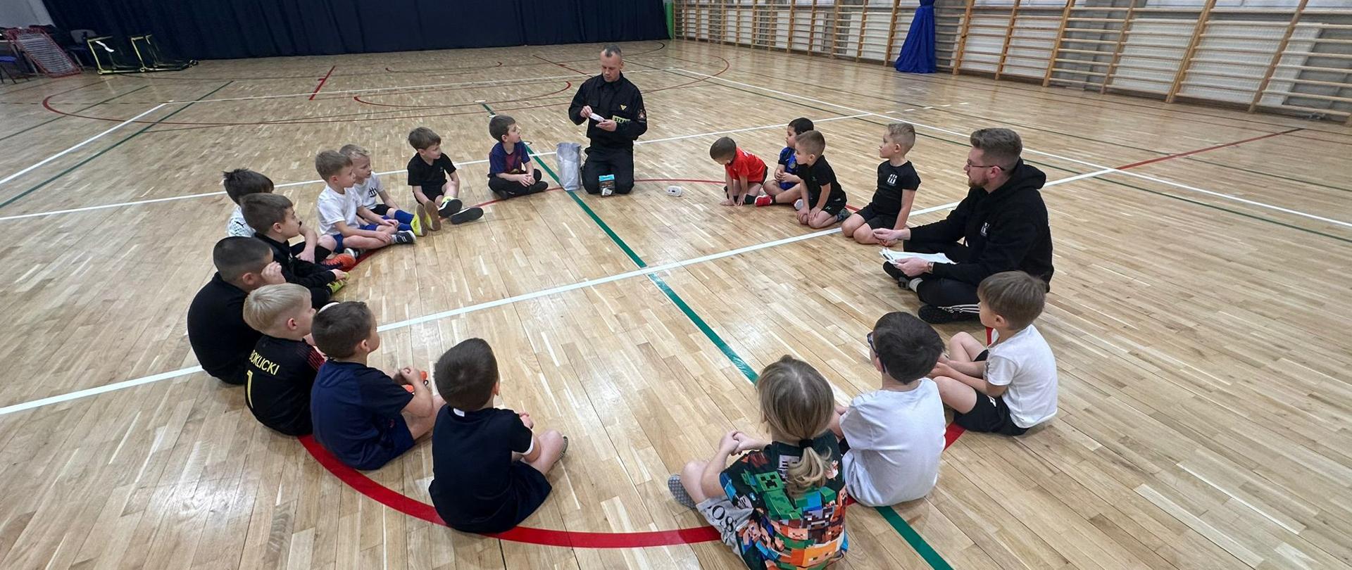 Pogadanka młodych zawodników ze strażakiem na temat zagrożeń mogące wystąpić zarówno w domu, jak i w codziennym życiu. Dzieci siedzą na parkiecie hali sportowej w kółku wysłuchując porad strażaka.