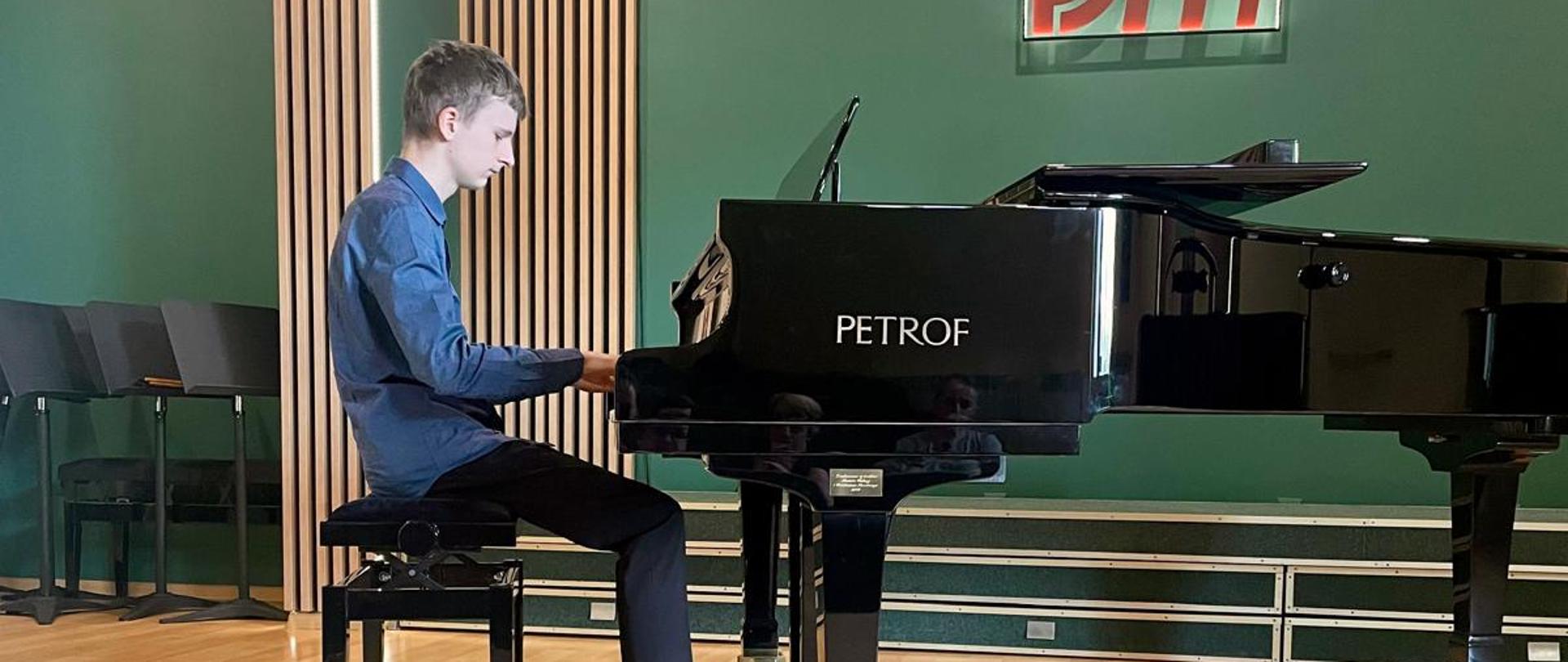 Na zdjęciu widać chłopca grającego na fortepianie. Jest ubrany w niebieską koszulę i ciemne spodnie. W tle znajduje się fortepian marki PETROF. Pomieszczenie ma nowoczesny wystrój, z drewnianą podłogą i zielonymi ścianami. Atmosfera sugeruje, że to występ lub próba.
