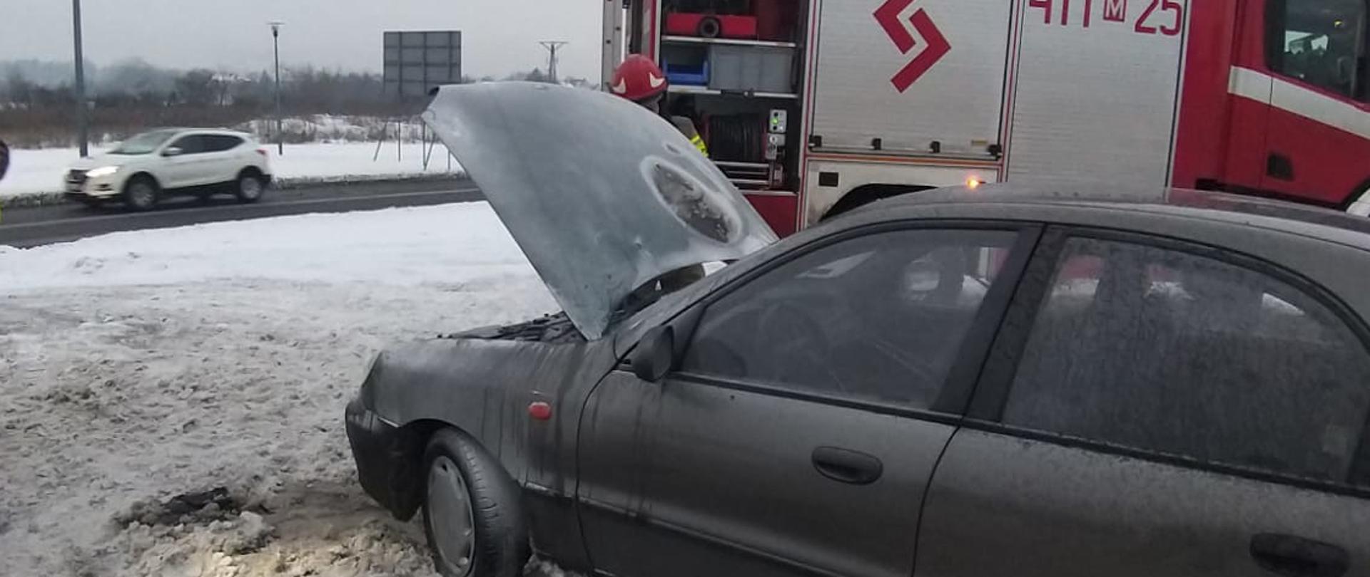 Na zdjęciu po pożarze samochód osobowy z podniesioną maską. Widoczny jest również samochód ratowniczo- gaśniczy straży pożarnej