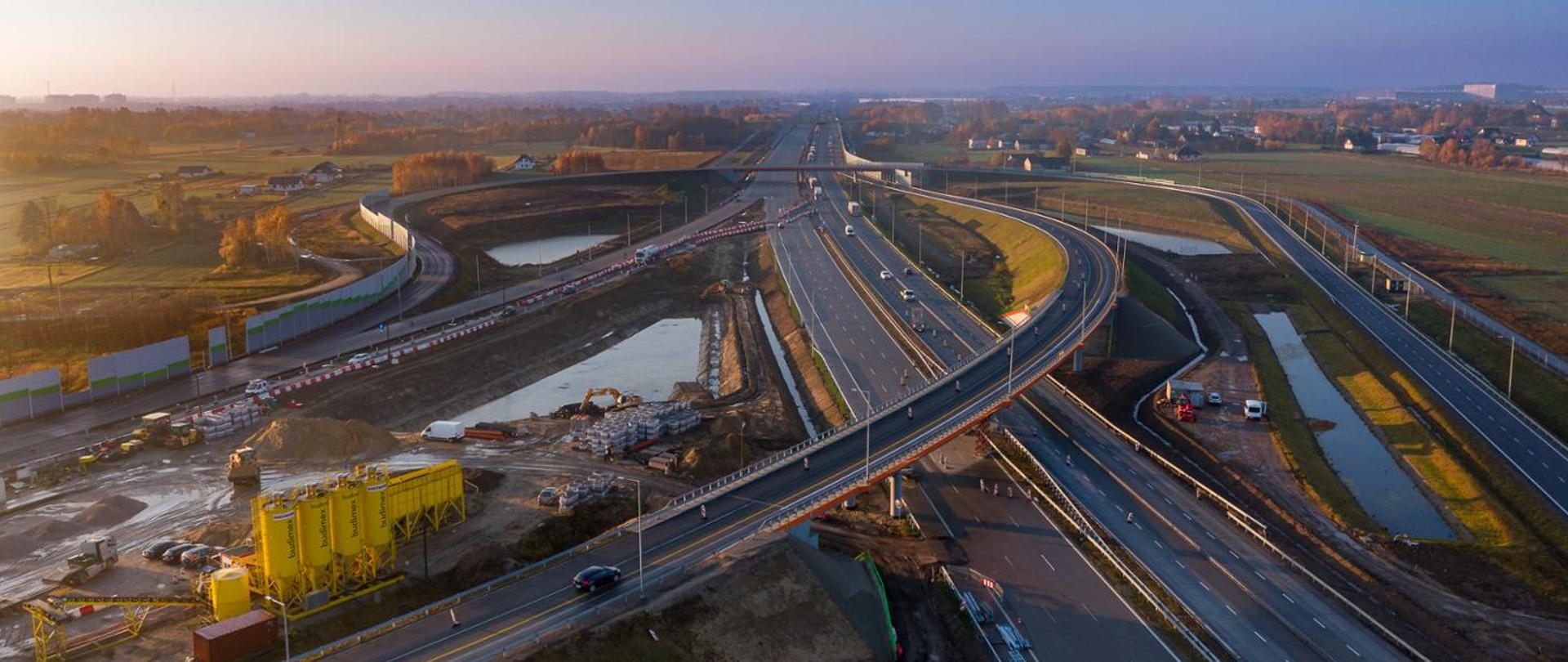 Widok na węzeł w porannym słońcu. Na pierwszym planie widać łącznicę węzła Warszawa - Katowice. Po prawej stronie zdjęcia, nowa łącznica dla kierunku Łódź - Warszawa. W lewym górnym rogu fotografii zaplecze budowy z żółtymi silosami.