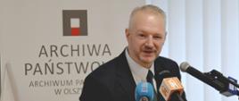 Wojewoda stoi przy mównicy na tle ścianki z napisem Archiwum Państwowe