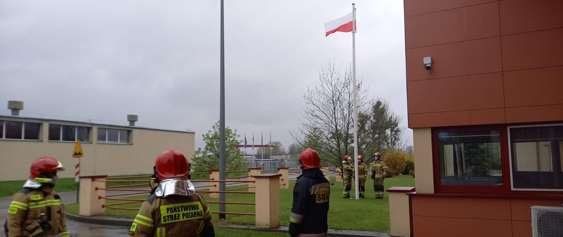 Zdjęcie przedstawia moment uroczystego podniesienia flagi narodowej na maszt przed obiektem Komendy Powiatowej PSP w Międzyrzeczu w dniu 2 maja 2021 r. przez strażaków z JRG Międzyrzecz.