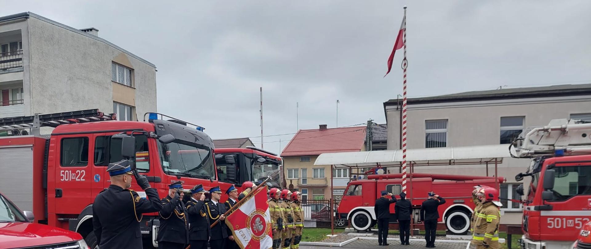 Funkcjonariusze Komendy, w tym poczet sztandarowy podczas uroczystej zmiany służby stoją na placu Komendy na przeciwko siebie. Po środku stoi Zastępca Komendanta Powiatowego. Poczet flagowy wciąga flagę na maszt.