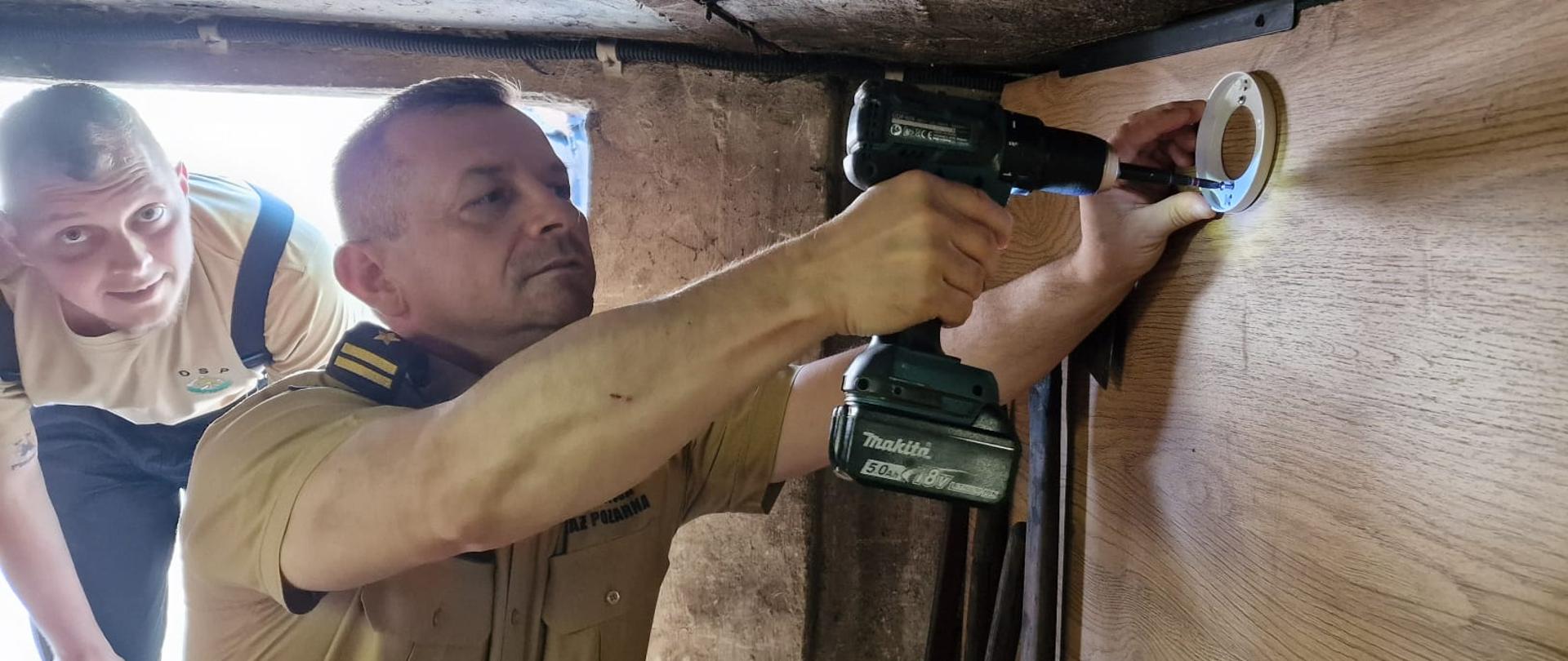 strażak przykręca wiertrką czujkę w mieszkaniu, za nim pochyla się strażak OSP