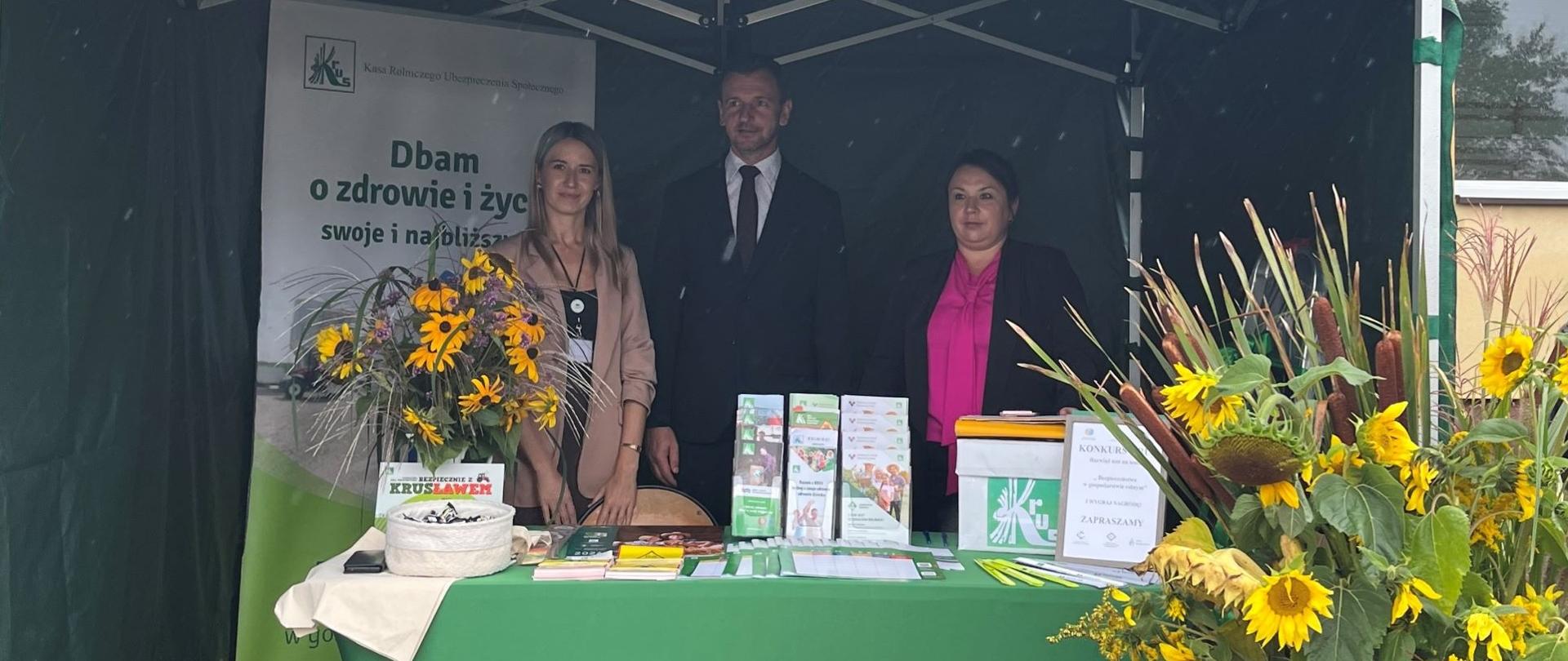 Przy stoisku informacyjno-promocyjnym Kasy stoją dwie kobiety i mężczyzna. Na stoliku przed nimi znajdują się materiały prewencyjne Kasy (ulotki, odblaski, plany lekcji dla dzieci).