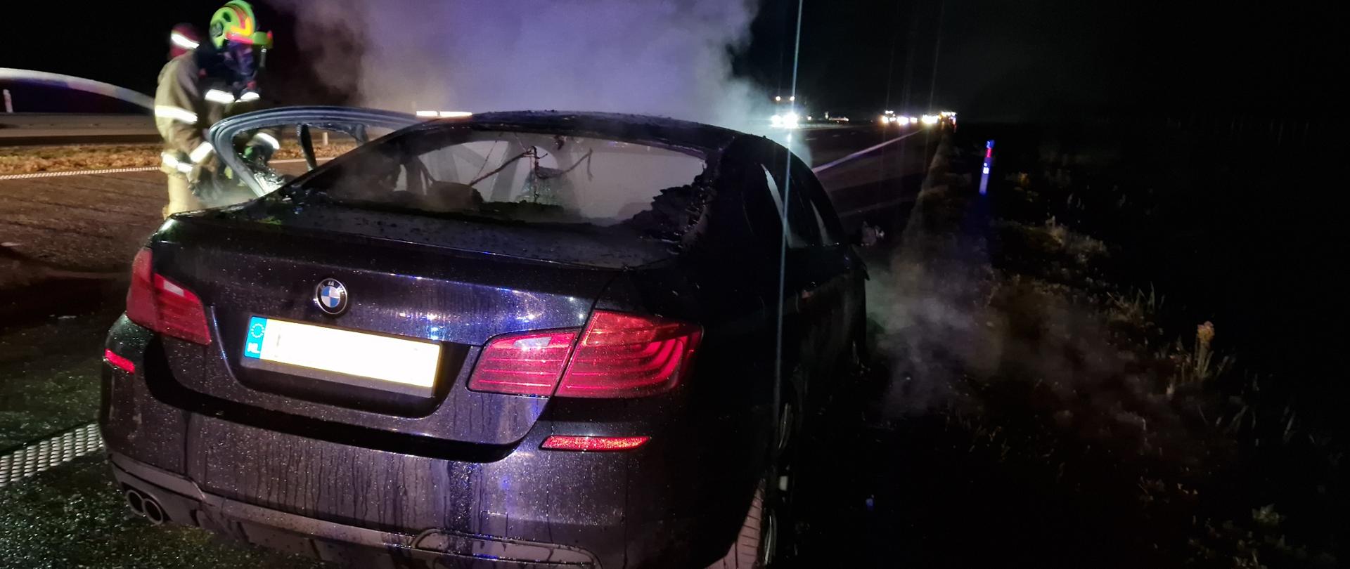 Na zdjęciu widać tył samochodu osobowego, wydobywający się dym z pod maski samochodu i strażaka w aparacie ochrony dróg oddechowych gaszącego samochód.
