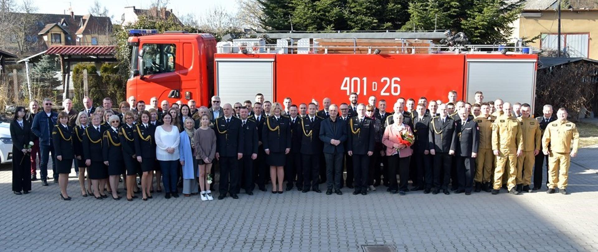 Zdjęcie przedstawia bardzo dużą grupę (kilkadziesiąt osób) uczestniczącą w oficjalnym spotkaniu lub uroczystości, pozującą na wybrukowanym placu. Tłem dla zgromadzonych jest bok dużego, czerwonego wozu strażackiego z widocznym numerem operacyjnym „401[D]26”. Po lewej stronie stoją głównie kobiety i mężczyźni w ciemnogranatowych mundurach wyjściowych ze sznurami galowymi oraz osoby w strojach cywilnych. Po prawej stronie szyku stoją strażacy w piaskowo-brązowych mundurach służbowych. Jedna z kobiet w centrum trzyma bukiet kwiatów. Scena rozgrywa się w słoneczny dzień przed budynkami jednostki.